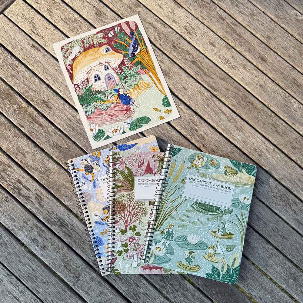 Three spiral-bound notebooks and an art print with whimsical forest and woodland illustrations are arranged on a weathered wooden plank surface.